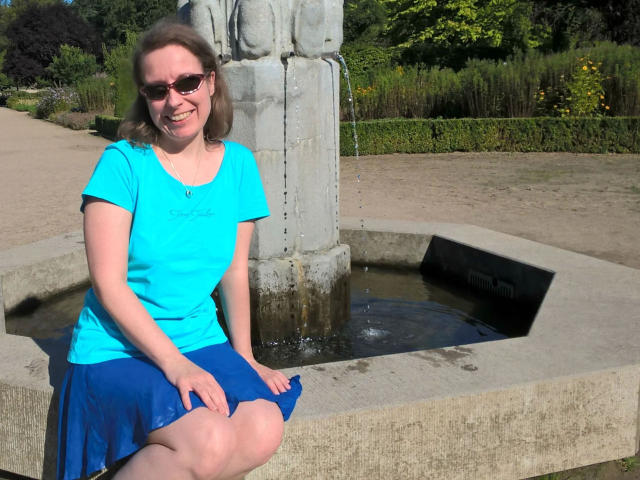 SexyAmyX-hot sitting by a fountain, wearing a blue shirt and skirt, with her ass slightly exposed, sunglasses on, smiling, and her hands on her thighs.