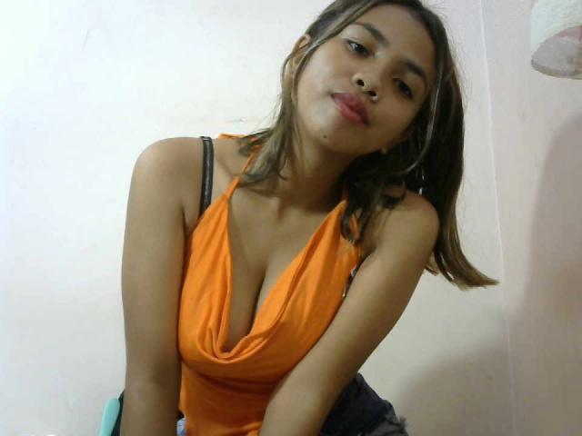 Merliah with light brown skin and straight brown hair, wearing an orange top with a deep cleavage, showing her small to medium-sized tits, and a black strap.