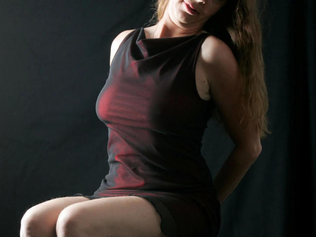 A photo of monelive-hot with long brown hair, wearing a tight black and red dress, sitting with hands behind her back, showing her ass and slightly visible tits.