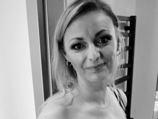 Black-and-white selfie of VanessaCute-hot with wavy blonde hair, subtle smile, dark eye makeup, small hoop earrings, and a delicate necklace, showing her bare shoulders.