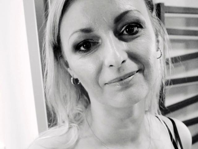 Selfie en blanco y negro de Vanessa. Es linda y atractiva, mostrando su rostro sonriente con labios brillantes, pequeños aros y cabello rubio ondulado, vistiendo una camiseta negra con tirantes. Sus ojos son oscuros y con un maquillaje ahumado..