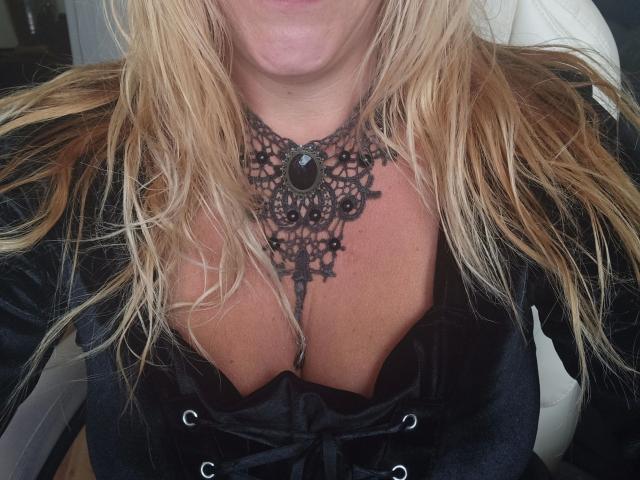 Femme aux cheveux blonds, portant un collier en dentelle noire, un corset à lacets noirs et une veste en velours noir, montrant ses seins et une pierre noire dans le collier..