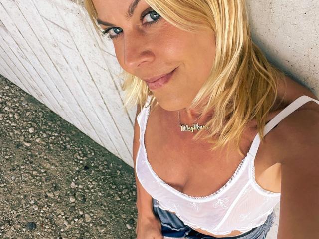Miley-Weasel with blonde hair, white sheer top, small tits, blue eyes, and a "Miley" necklace, sitting on gravel, smiling at the camera.