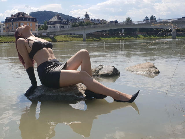 FetishQueenofPain-hot with red hair, black bra, black skirt, black heels, and black gloves, sitting on a rock in a river, legs crossed, head tilted back, water reflecting.