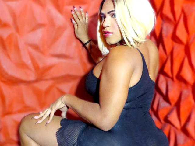 ZahorySexy with blonde hair, wearing a black dress, showing her ass, back to the camera, hands on a red textured wall, looking over her shoulder with red lipstick.