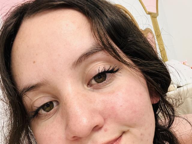 Le visage de ChloeDulce, avec ses yeux marron et ses longues cils noirs, sa peau légèrement rosée et ses cheveux ondulés foncés, un petit sourire et quelques taches de rousseur..