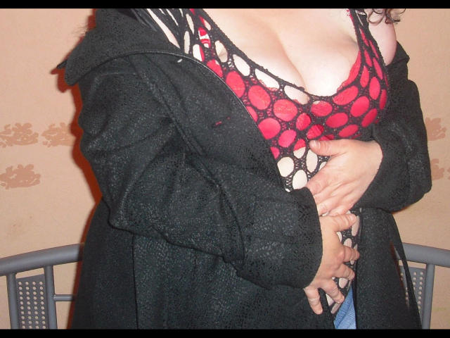 REALAMATEUR-hot with large boobs in a red and black mesh bra, covered by a black jacket, holding her tits with her hands. Beige wall and chair in background.