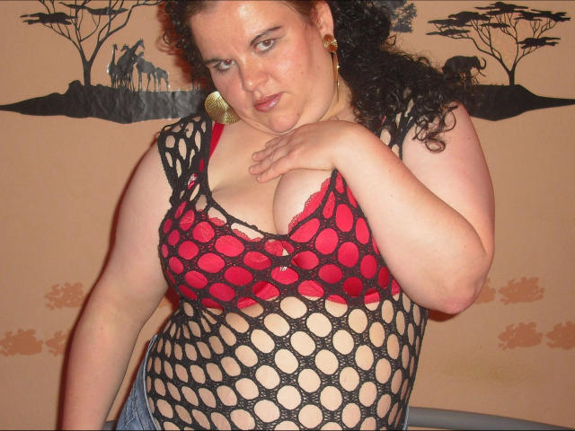 REALAMATEUR-hot with curly brown hair, wearing a black fishnet top over red bra, covering her big tits with one hand, looking down with a slight smirk.
