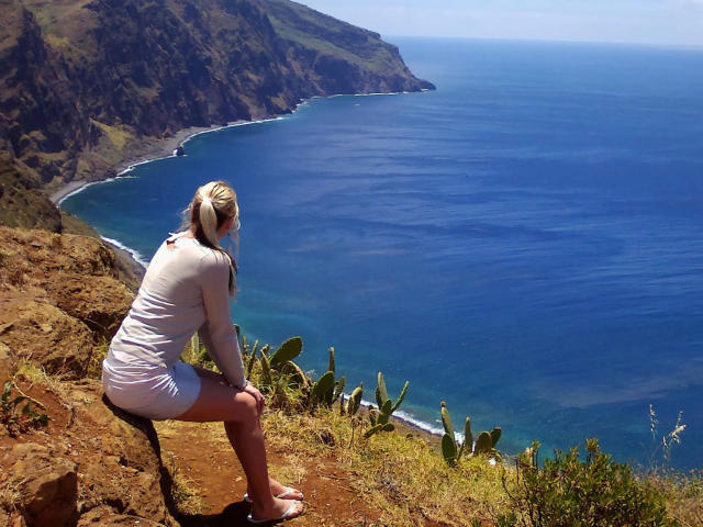 SeiMeinVoyeur: una donna con una coda di cavallo bionda, un vestito bianco, seduta a gambe accovacciate su una scogliera rocciosa, con l'oceano blu, le colline verdi e il cielo sereno, che guarda l'acqua, con i glutei leggermente sollevati..