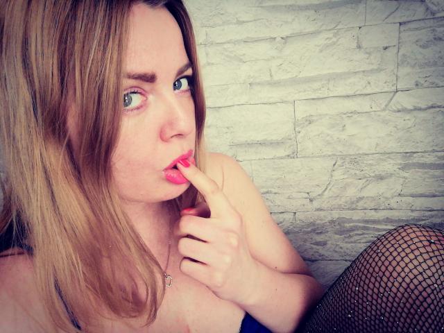 SeiMeinVoyeur - uma mulher atraente, com cabelo loiro, olhos azuis e batom rosa, chupando o dedo, vestindo meias de rede, mostrando o ombro nu e um colar, com uma aparência sedutora..