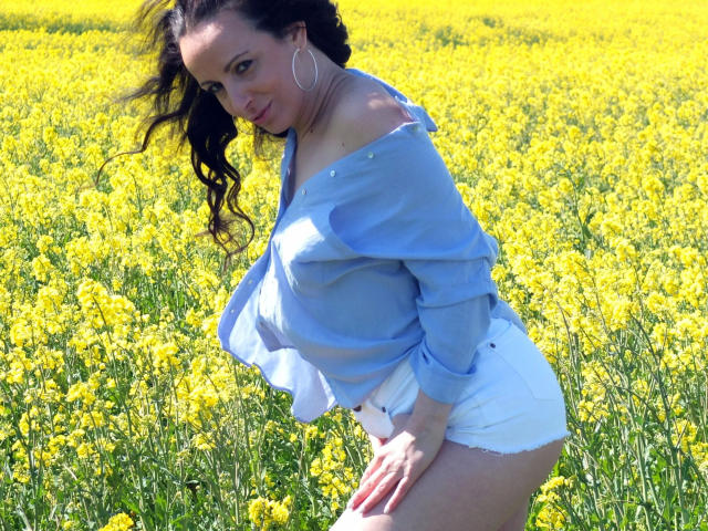 LuderMeggie-hot has dark curly hair, blue off-shoulder shirt, white shorts, and large hoop earrings, standing in a yellow field, leaning forward with a playful smile.