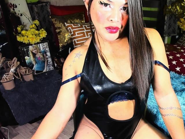 TransGiftedCum in a shiny black cutout one-piece, sitting with long brown hair, red lipstick, star tattoos, and a sexy gaze, showing their small tits and blue lace straps.