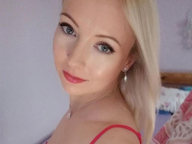 SusiPrivat224-hot with blonde hair, blue eyes, red lipstick, and pink straps, smiling at the camera. She's wearing earrings and a necklace. Her tits are slightly visible.