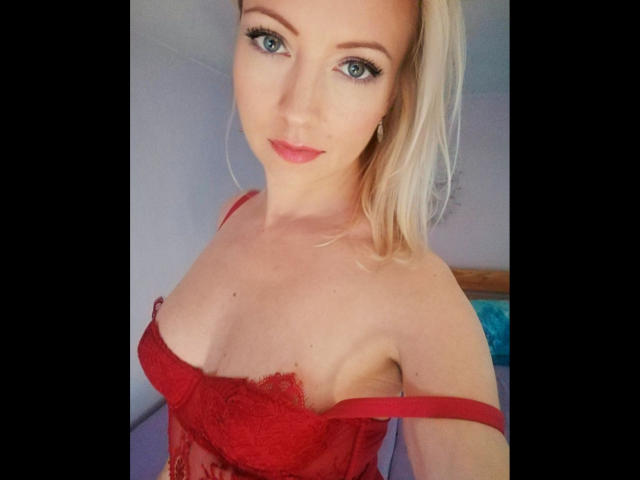 SusiPrivat224, mit blonden Haaren, blauen Augen und rotem Lippenstift, trägt einen roten Spitzen-BH, wobei der rechte Träger heruntergerutscht ist und ihre Brüste sichtbar sind. Sie schaut in die Kamera..