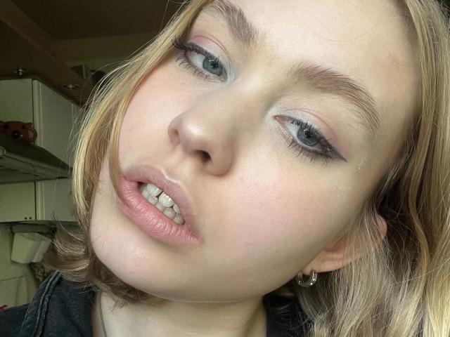 BrandyHelga-hot's face, showing pale skin, blue eyes with light makeup, slightly parted pink lips, and blonde hair, looking up with a relaxed expression. Small silver hoop earrings.