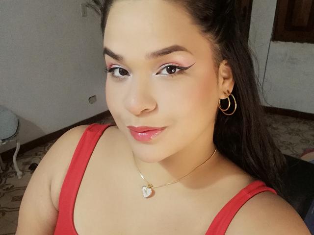AntonelaPark with shiny pink eyeliner, red lipstick, and glossy lips, wearing a red tank top, gold hoop earrings, and a heart necklace, smiling at the camera.
