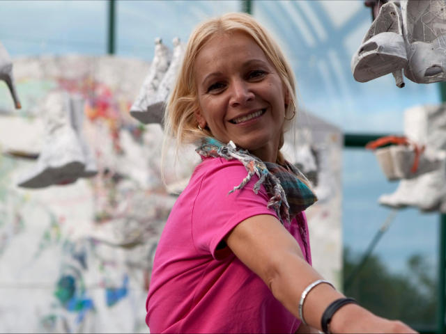 Lulu-4-you, blonde woman, pink shirt, smiling, holding up white sneakers, colorful scarf, blue bracelet, outside, bright day, light brown skin, mid-40s, casual, relaxed, happy.