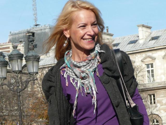Lulu-4-you has blonde hair, a purple top, gray cardigan, colorful scarf, and black bag. She's smiling with wind in her hair. Her tits are small, and she's cute.