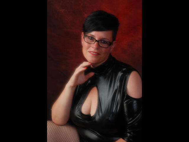 Molly-Sun with short black hair, glasses, and a shiny black latex dress showing her big boobs and cleavage, sitting against a dark red background, smiling slightly.