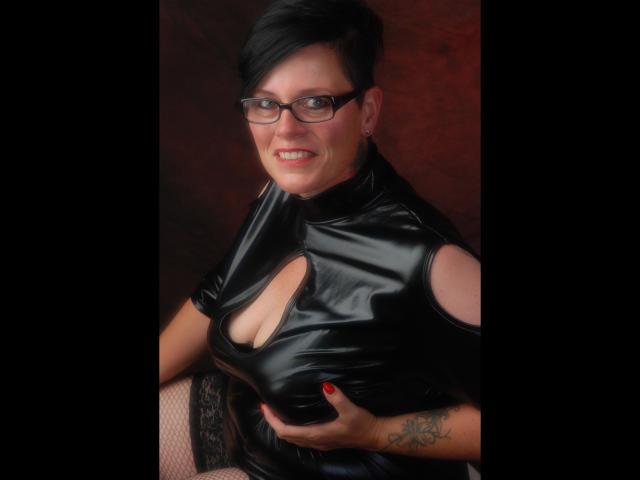 Molly-Sun with black hair, glasses, and red nails, wearing a shiny black latex top with a cutout, holding her tits, showing a tattoo on her right hand.