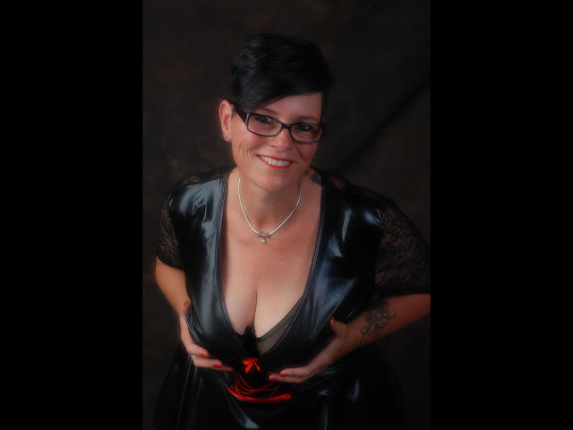 Molly-Sun with glasses, black hair, smiling, wearing a shiny black dress with lace sleeves, holding her tits, red ribbon at bottom, necklace, tattoo on right wrist.