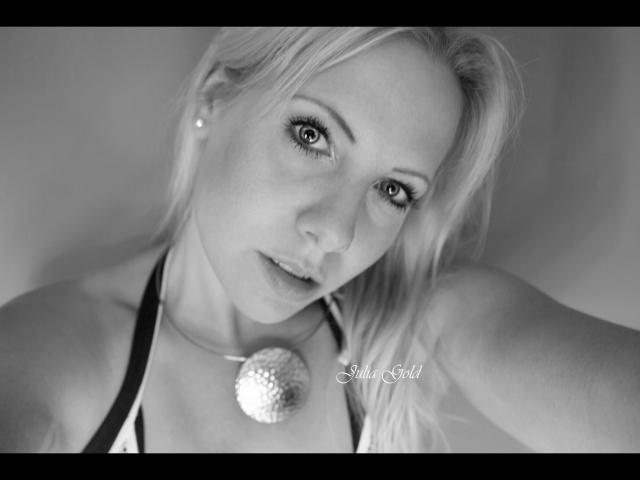 JuliaGold-hot tiene el cabello rubio, un arete de perla y un collar redondo y texturizado. Está mirando a la cámara con ojos grandes, una nariz pequeña y una sonrisa sutil..