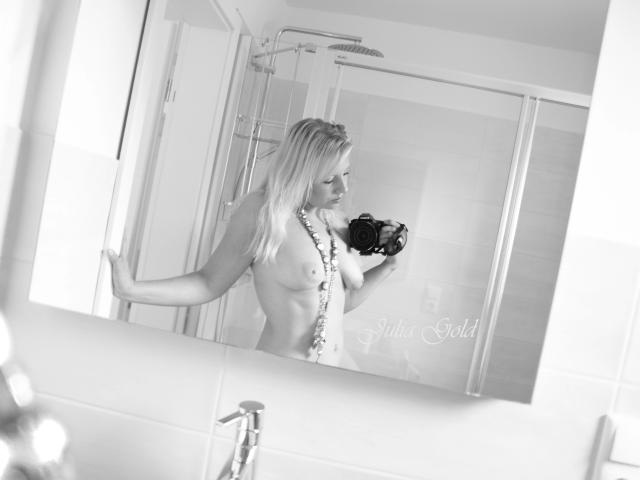 Fotografía en blanco y negro de JuliaGold, sin ropa, con senos pequeños, sosteniendo una cámara, con un collar largo, de pie frente a un espejo que la refleja, en un baño moderno con ducha de vidrio..