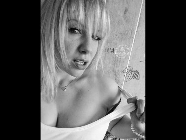 JuliaGold, de cabello rubio, con un guiño, sosteniendo un abanico, bajándose una camiseta sin mangas para revelar sus grandes senos desnudos. Blanco y negro, collar, labios entreabiertos..