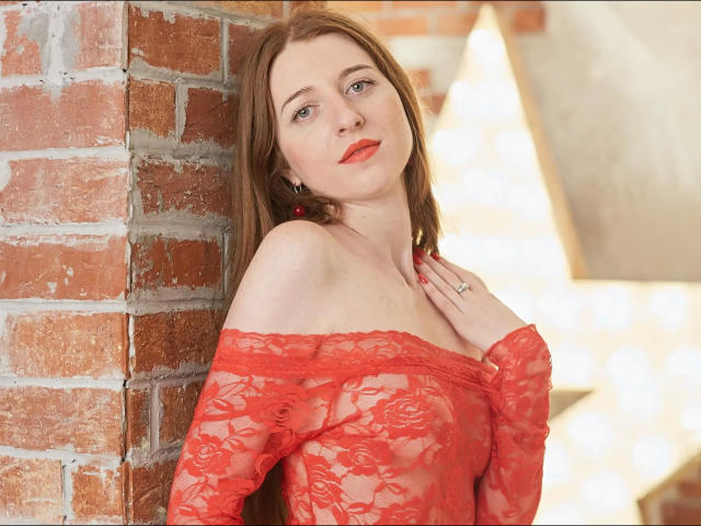 Sweetyangel27, con una blusa roja de encaje que se lleva sin hombros, mostrando sus pequeños pechos, apoyada contra una pared de ladrillo, mirando a la cámara con lápiz labial rojo y con una mano en el pecho..
