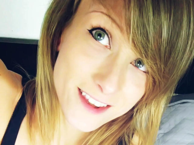 AliceKinkycat-hot has blonde hair, green eyes, and a cute smile with pink lips, wearing a black top, in a selfie with slightly messy hair.