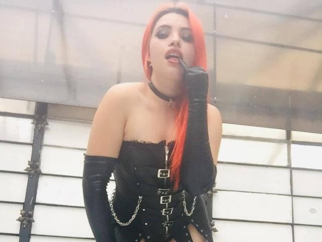 ElizaLovely met rood haar, een zwarte choker, een behaalde corset zonder bandjes en zwarte handschoenen, die een sigaret aan haar mond houdt, en verleidelijk kijkt met haar borsten die licht zichtbaar zijn..