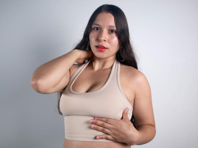 AnneQueen met lang bruin haar, rode lippenstift, een beige geribbelde tanktop, met haar hand in de buurt van haar nek, de andere hand op haar buik, waarbij haar borsten zichtbaar zijn, lichte huid, lichtelijk rond, staand..