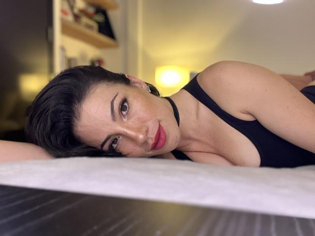 Liliysweet is lying on a bed, head resting on her arm, wearing a black tank top, with short black hair and red lipstick, looking at the camera.