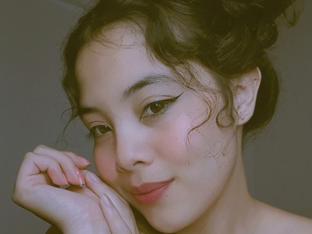 KileyDorn has wavy brown hair, black winged eyeliner, pink lipstick, and is gently touching her chin with pink-manicured nails, smiling softly. Her skin is light and she's cute.