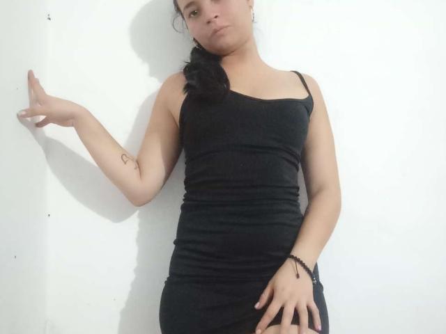 LiliHotter, wearing a black tank dress, has her right arm raised, showing a small tattoo on her inner wrist, and her left hand on her thigh, looking annoyed.
