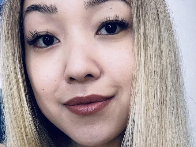 RenyLime's face, blonde hair, brown eyes, full lips with dark red lipstick, small moles on her cheek, thick black eyelashes, slight smile, fair skin, no visible makeup.