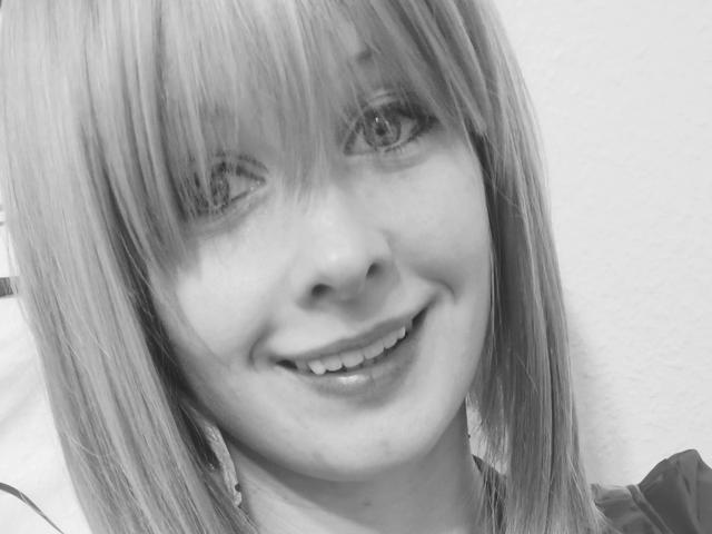 Black-and-white selfie of annekehot-sex with blonde hair, bangs, smiling, showing teeth, and wearing a shirt. Her eyes are slightly winking. Her nose is small and her lips are glossy.
