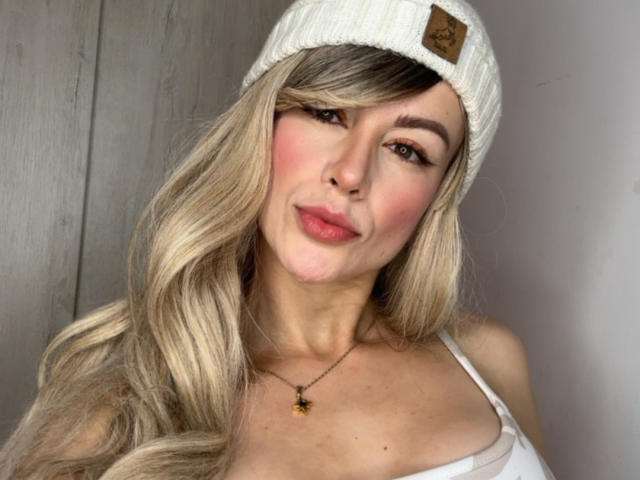 Leared has long, wavy blonde hair, wearing a white beanie and a white tank top, with pink blush on her cheeks, and a necklace with a small yellow pendant.