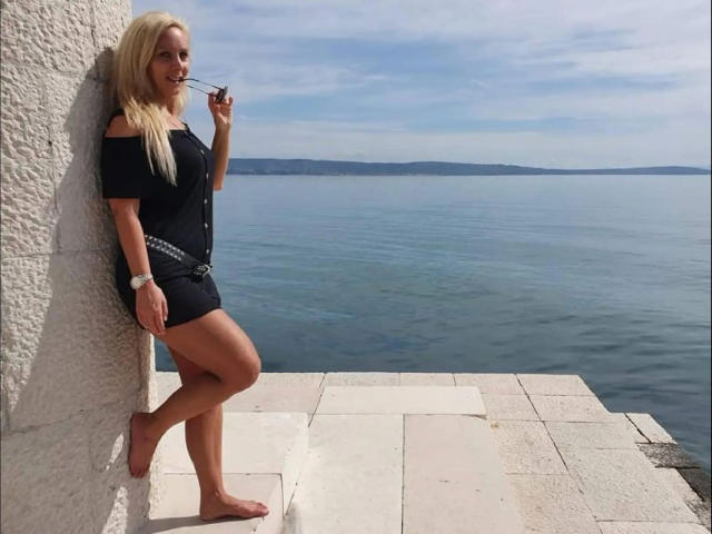 Annabel-Massina leans against a white wall, barefoot, wearing a black off-shoulder dress, holding a cigarette, with a blue sea and cloudy sky in the background.
