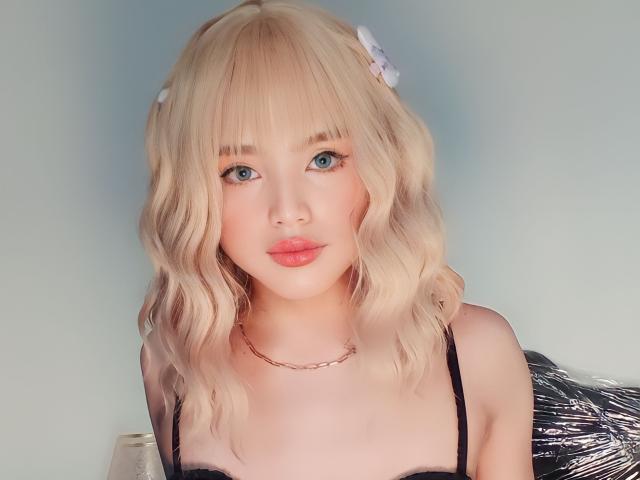 Redhuxley heeft blond, golvend haar, blauwe ogen, roze lippen en draagt een zwarte top met bandjes en een gouden ketting. Ze kijkt naar de camera met een fronsende uitdrukking..