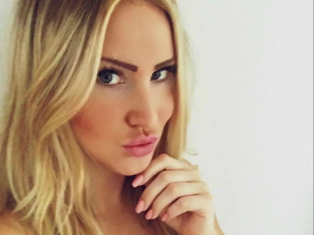 Blonde Fitness-Maus with wavy hair, green eyes, and pink lips, looking seductively at the camera, hand near mouth, pink nails, white background.