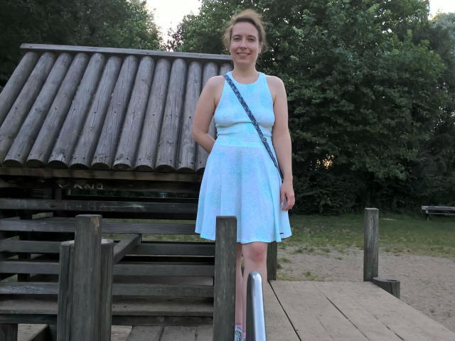 SexyAmyX-hot in a light blue dress, standing on a wooden dock, smiling, with a blue strap bag, wooden roof structure, and green trees in the background.