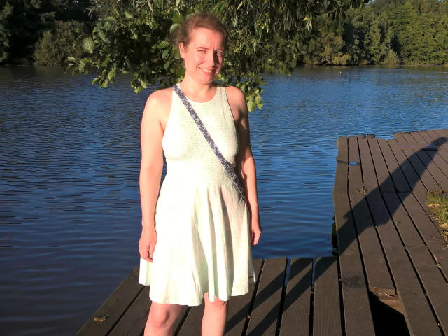 SexyAmyX-hot with pale skin, short brown hair, small tits, white sleeveless dress, standing on wooden dock, smiling, blue water, green leaves in background, daytime, natural light.