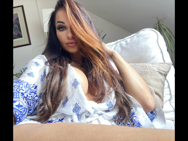 SexyStacyS-hot with long brown and blonde hair, blue patterned top, exposing large tits, sitting on a white couch, hand in hair, green eyes, looking at camera.