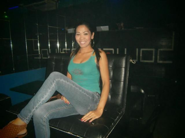 xUrDestinyTS69x-hot with long black hair, green tank top, and blue jeans, sitting on a black leather chair, smiling, wearing brown shoes, in a dimly lit club.