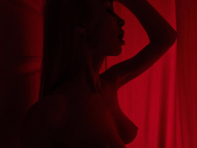 BlondiAngel-hot's silhouette in red light, raising arm, with visible tits, long hair, and a sexy profile, looking to the side. Red curtain in background.