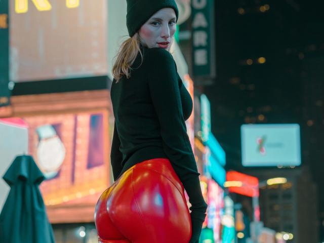 HannaSecret-hot with blonde pigtails, black beanie, black top, and shiny red butt-flesh pants, looking back with a slight smirk in a neon-lit urban night setting.