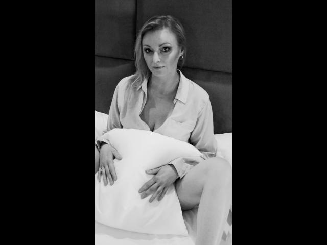 VanessaCute-hot with blonde hair, wearing an unbuttoned shirt, holding a pillow, sitting on a bed, showing her tits, looking serious, in a black-and-white photo.