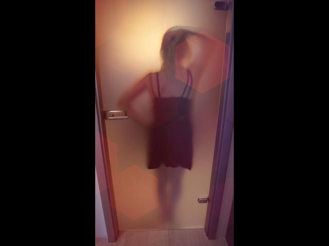 Vanessa : Mignonne et séduisante, avec une silhouette floue et brumeuse, debout dans un encadrement de porte, vêtue d'une robe sombre, et tenant une sangle, dos à la caméra, avec une poignée en métal sur la gauche..