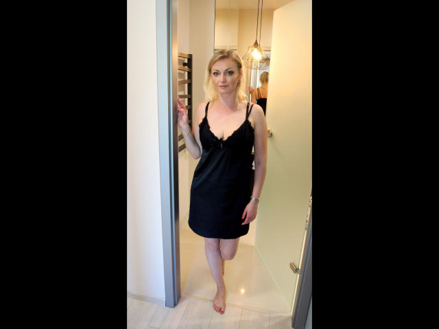 VanessaCute-hot está de pie en un umbral, vestida con un vestido negro, sin mangas, con encaje y escote bajo, que muestra sus pequeños pechos y su trasero desnudo, descalza, con cabello rubio..